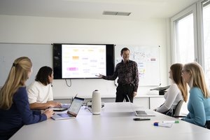 Personen am Tisch, eine Person vor einem digitalen Whiteboard