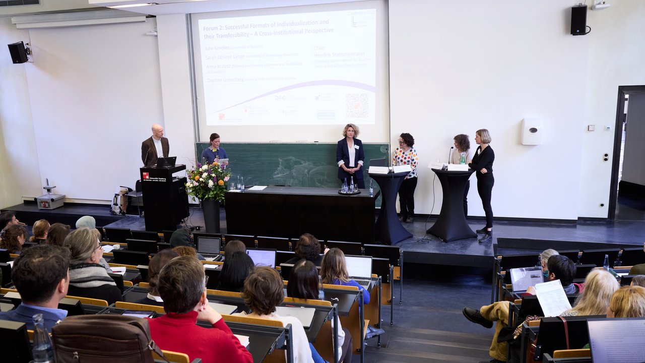 Blick aus den Sitzreihen eines Hörsaal auf das Podium mit Personen.