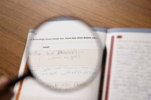 Lupe vor einer Seite aus einem Schulbuch mit handschriftlichen Einträgen