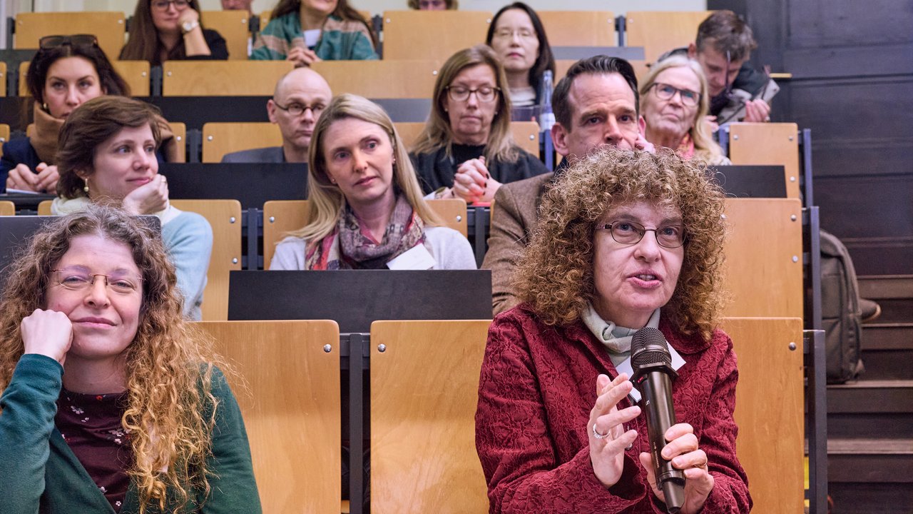 Zuhörende im Saal, eine Person vorne mit Mikrophon.